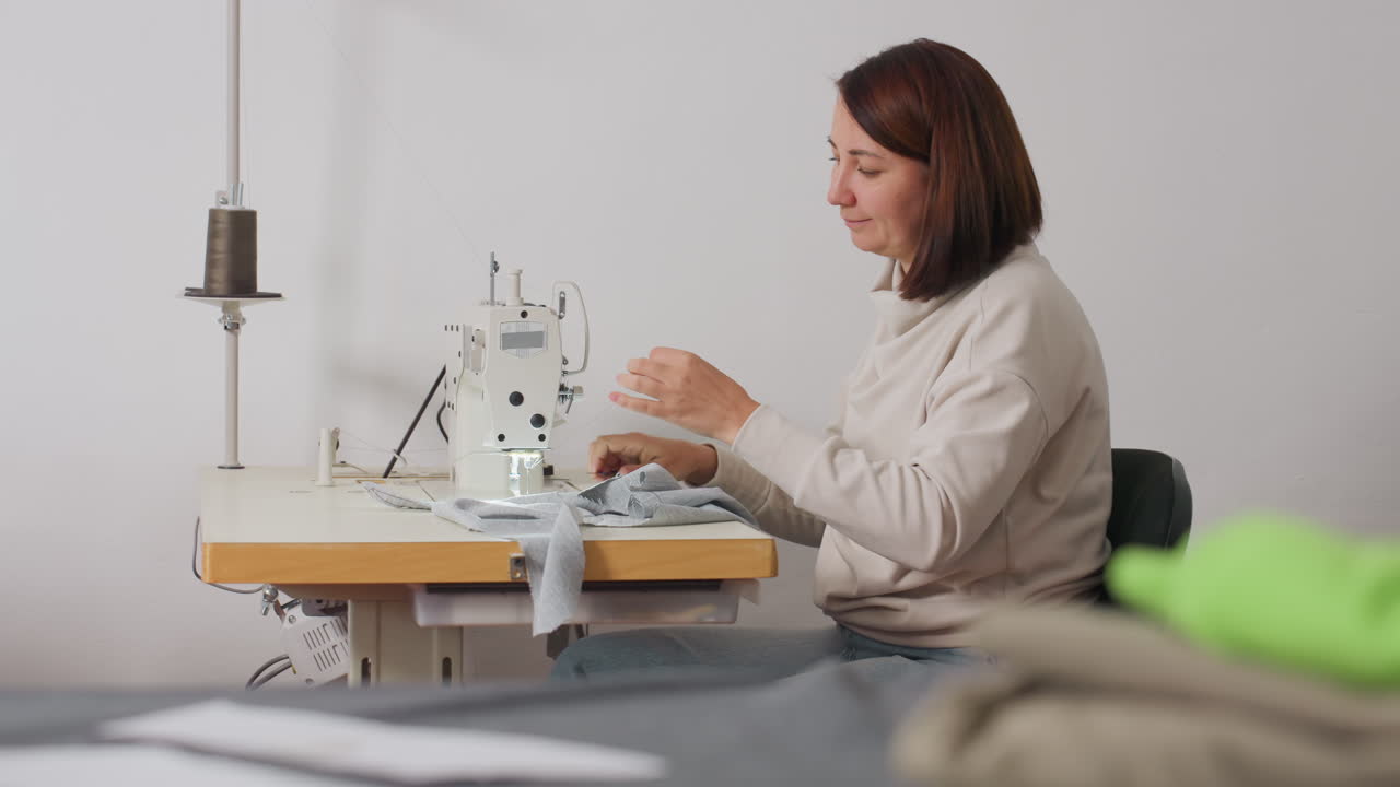 Clothing designer cutting tip of thread and reattaching it onto needle in sewing machine, smiling while adjusting stitches, demonstrating meticulous technique and glamorous vibe in minimal workspace
