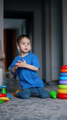 little kid wearing blue t-shirt and pants sits on the floor among his toys. toddler stands up and approaches the camera. vertical video.
