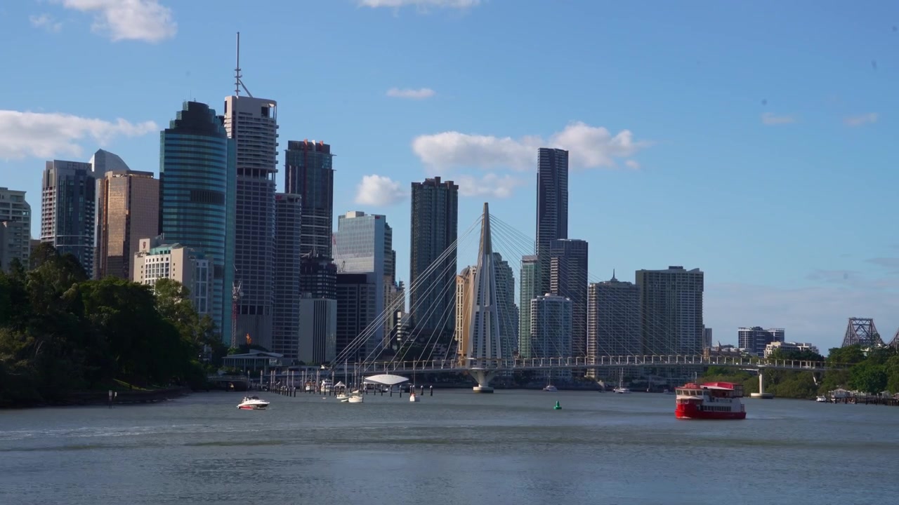 Ripresa in time-lapse dello skyline di Brisbane e del caratteristico Kangaroo Point Green Bridge che attraversa il fiume sotto un cielo azzurro e limpido, con traghetti che navigano sull'acqua.