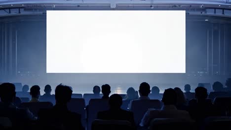 spectators sitting in an auditorium are focused on a blank white screen, creating a quiet atmosphere for presentations