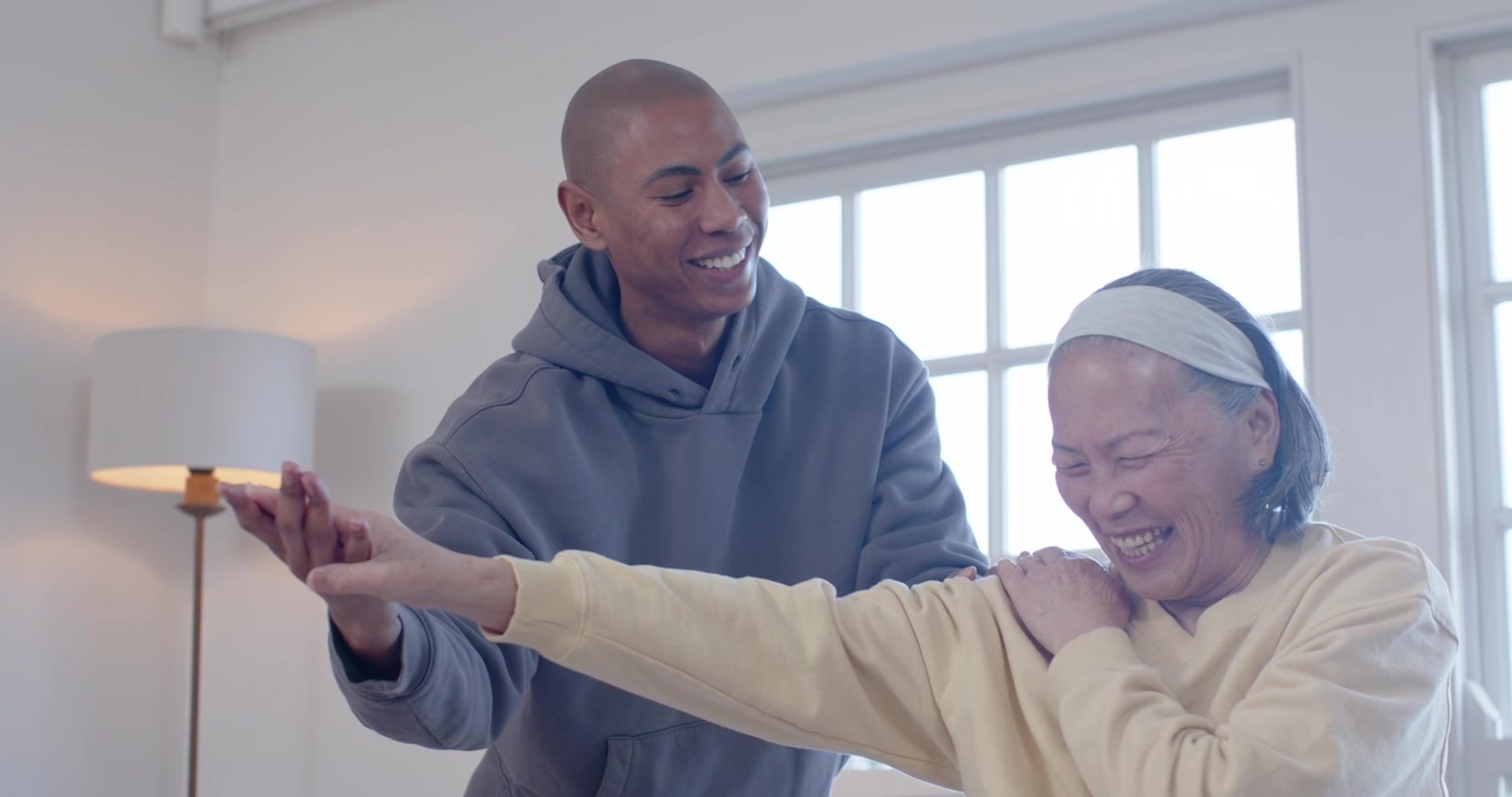 Senior Asian woman on ball while African American man guiding shoulder stretch for mobility at home