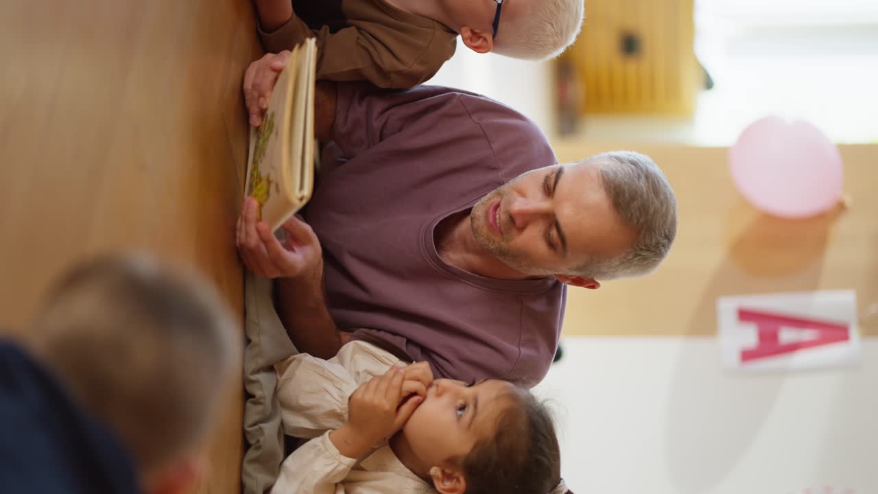 Vertical video of a man with gray hair in a purple T-shirt reading a book to children who are lying on the floor on special pillows and listening to him in a club for preparing children for school