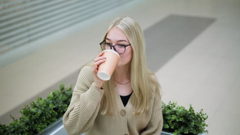 young lady seated in shopping mall sipping beverage while looking away, shoppers can be seen walking in the background with a blurred view of their legs and busy mall environment