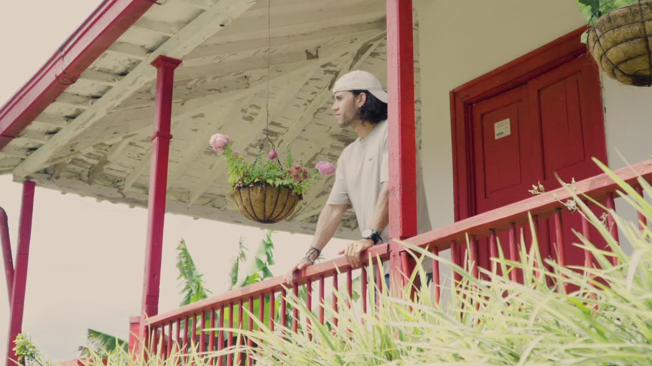 A man leans out a colorful balcony of a traditional house in Santa Bárbara, Antioquia, surrounded by vibrant plants.