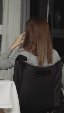 woman with disability sits in kitchen drinking water from glass. lonely young person looks out dark window sitting near table at home backside view