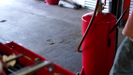 Mechanics-garage-slow-pan-up-from-red-tool-box-with-air-compressor-tools-and-oil-changing-equipment-in-focus,-as-mechanic-is-out-of-focus-in-background-working-on-car-on-lift