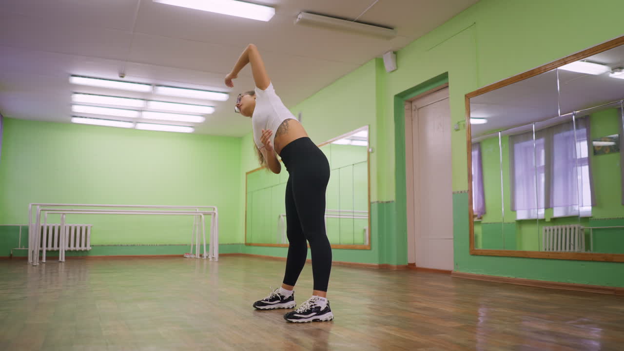 Front side view of girl in white top and black leggings stretching side to side in bright indoor studio with wooden floor, mirror wall, and green painted background, focusing on warmup