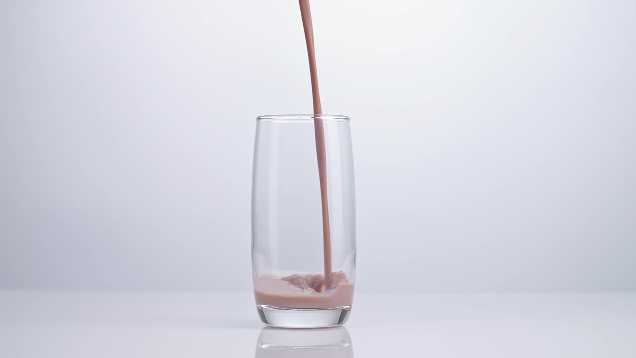 Versamento al rallentatore di bevanda al latte al cioccolato in un bicchiere tumbler su sfondo bianco