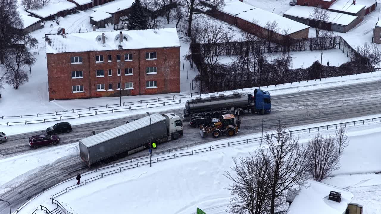 Aerial winter scene of a jackknifed semi trailer blocking an icy city road as cars pass and a recovery vehicle assists after a traffic accident in snowy conditions