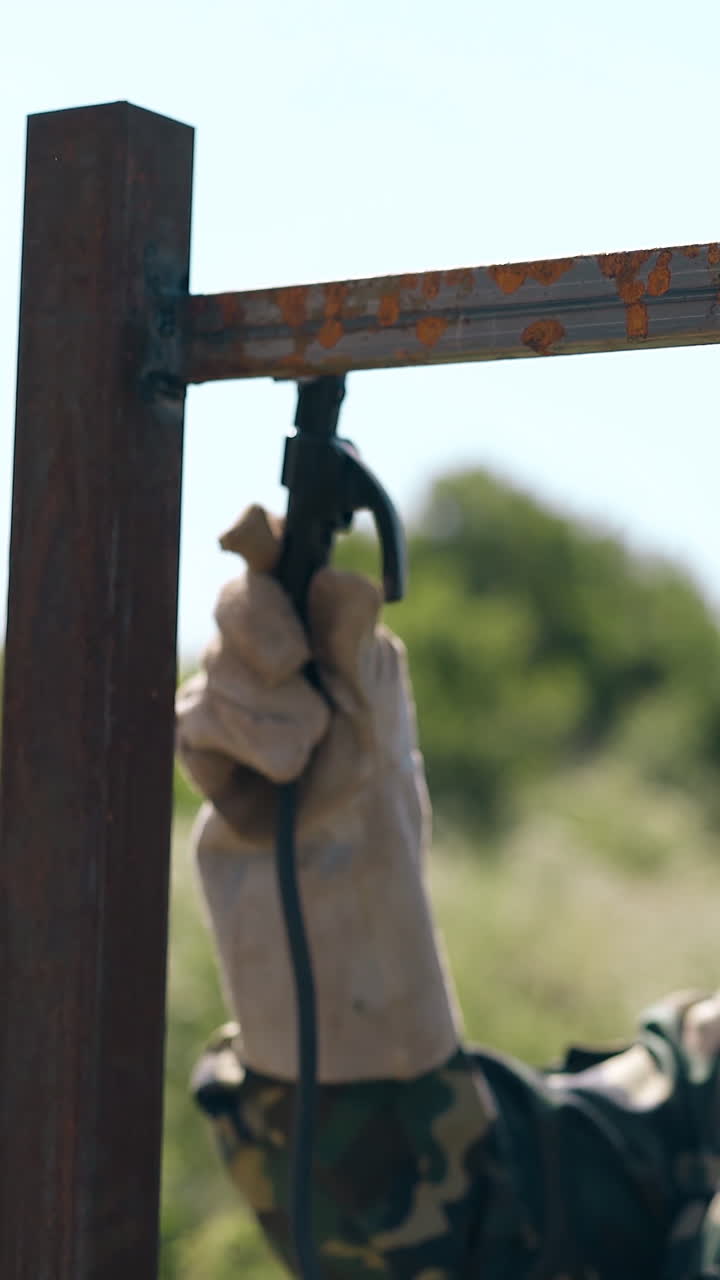 skilled builder in camouflage jacket welds upper plank of new fence with modern equipment on sunny day