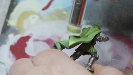 male hands painting miniature fantasy figure model green with flat paint brush and texture palette mat below with different colors