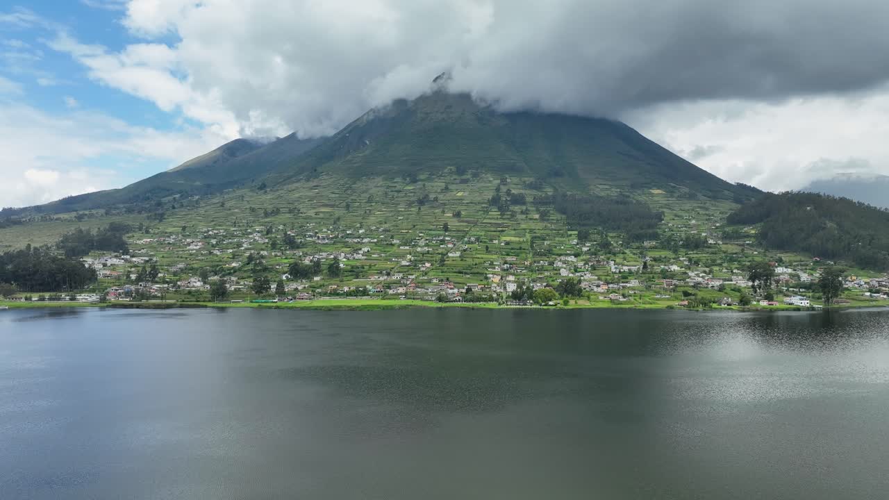 Green mountain top covered with fluffy clouds with a town at shore of a sea. Drone view.