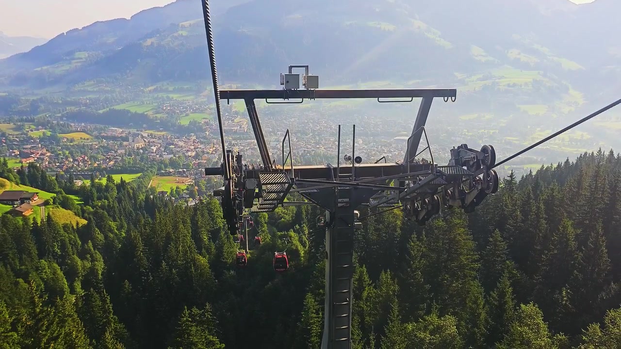 Gondole rosse salgono su un pendio boscoso circondato da prati alpini, chalet, strade tortuose e montagne panoramiche, catturando un pacifico paesaggio di una località di villeggiatura estiva in Europa.