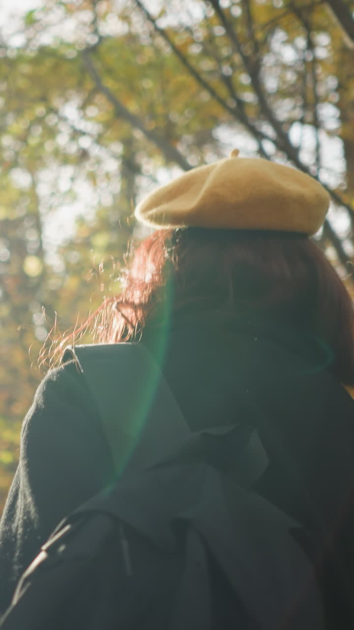 Lady walking through forest path adjusting bag with yellow beret and sunglasses as warm sunlight creates a glowing effect around her, enhancing the autumn atmosphere and peaceful nature scenery