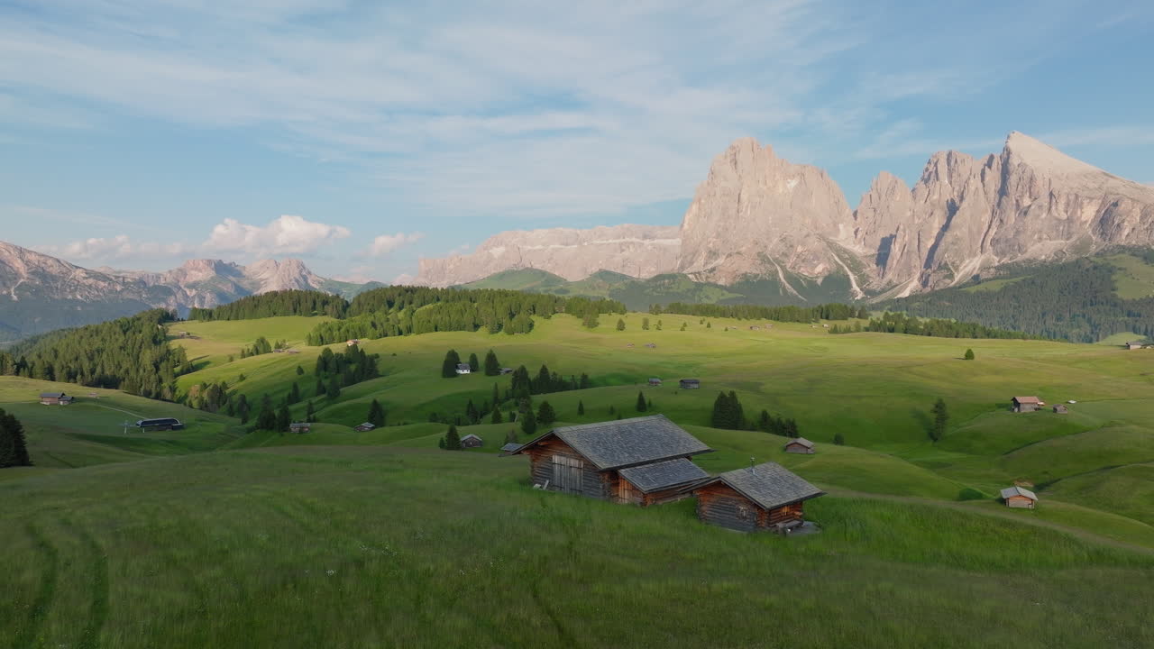 Slow panning drone shot of mountain huts in Alpe di Siusi during sunset. Vibrant grasslands and Dolomite peaks in the background