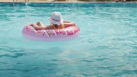 a funny little girl in a panama skates on an inflatable circle in a large pool in a hotel on the sea. rear view