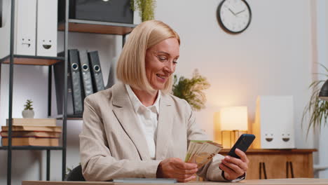 Mature-businesswoman-counting-money-euro-cash-using-smartphone-calculator-app-at-home-office-desk