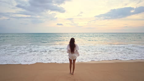 Una-Mujer-Joven-De-Espaldas-A-La-Cámara-Espera-A-Que-Suba-La-Marea-En-Una-Playa-De-Arena,-En-El-Sudeste-Asiático