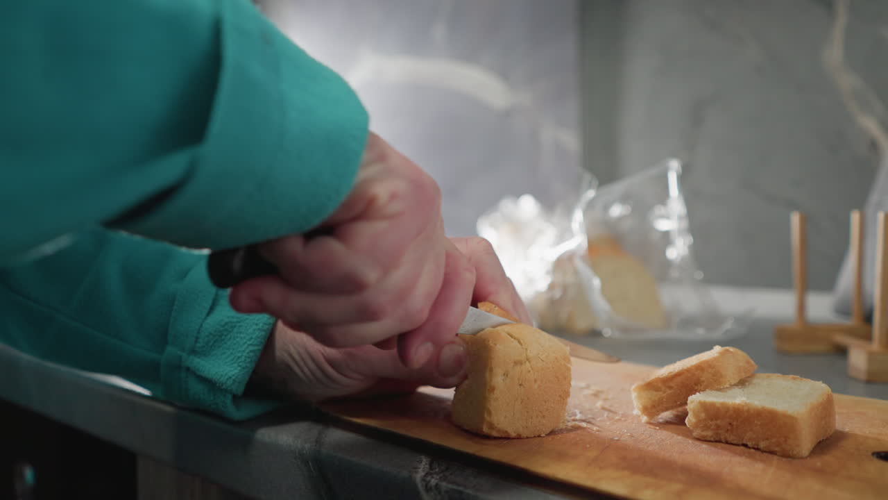 Close-up shot of older woman slicing freshly baked bread with a knife on wooden cutting board. Fresh bread slices and kitchen tools visible, soft lighting, and modern kitchen setting
