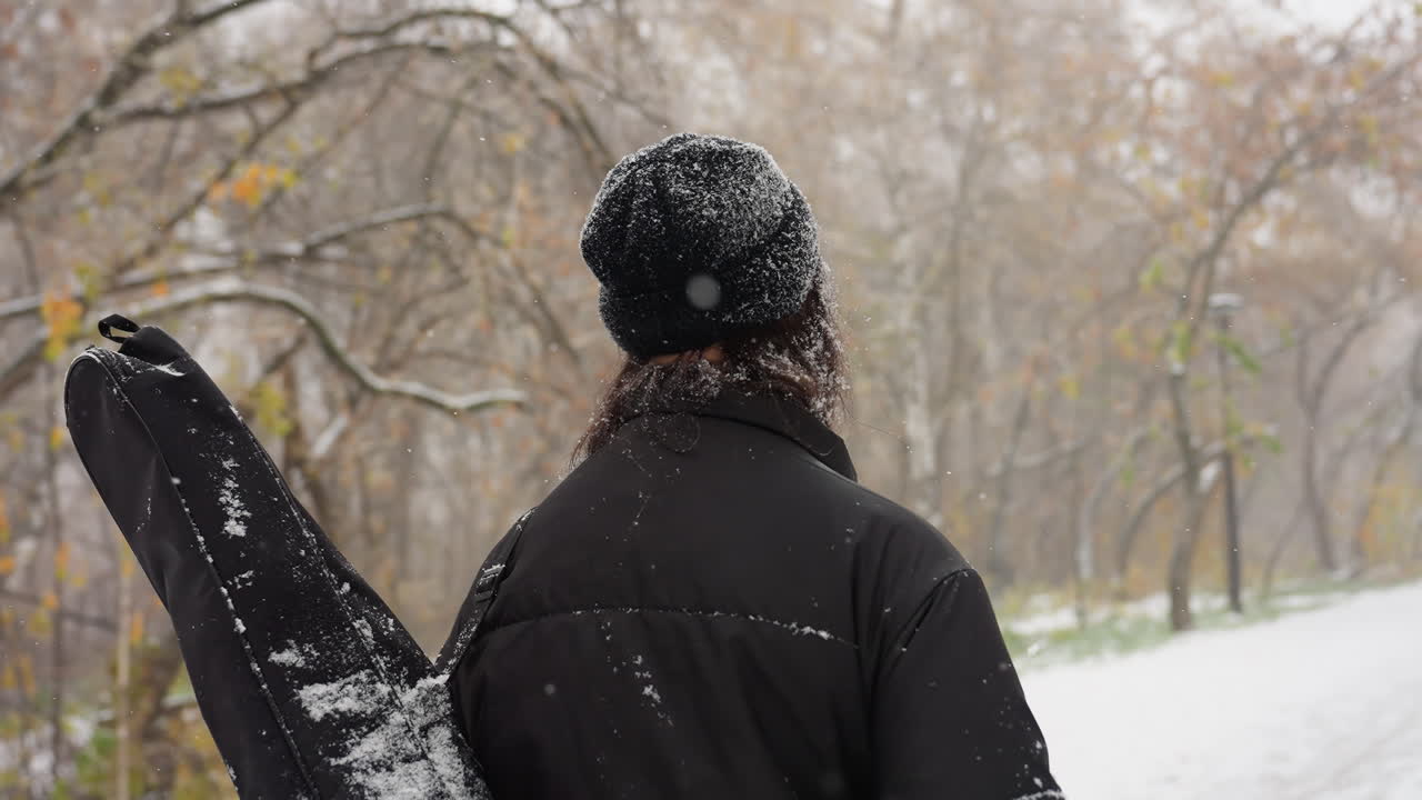 vista de fondo de una persona caminando por un camino nevado del parque, cargando un paquete de guitarra en el hombro, rodeada de árboles helados y un paisaje invernal sereno