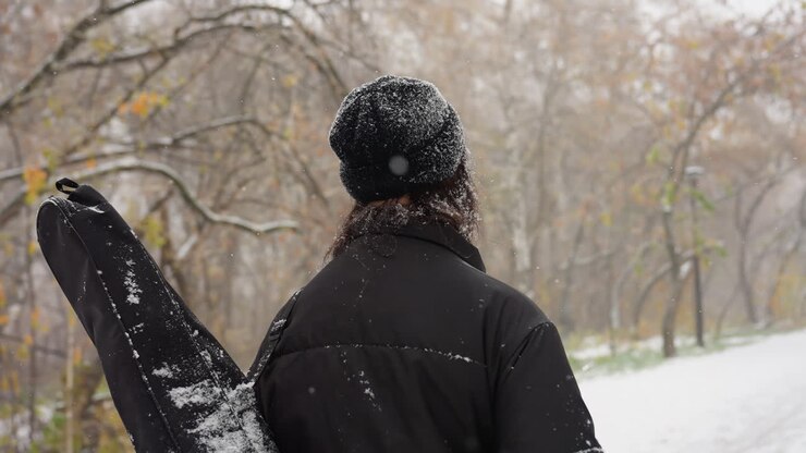 Primo piano della vista posteriore di una persona che cammina lungo un sentiero innevato del parco, con una custodia per chitarra in spalla, circondata da alberi ricoperti di brina e un sereno paesaggio invernale