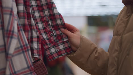 close-up shot of a hand wearing a light brown jacket, gently touching plaid shirts hanging on a clothing rack. the person's face is not visible