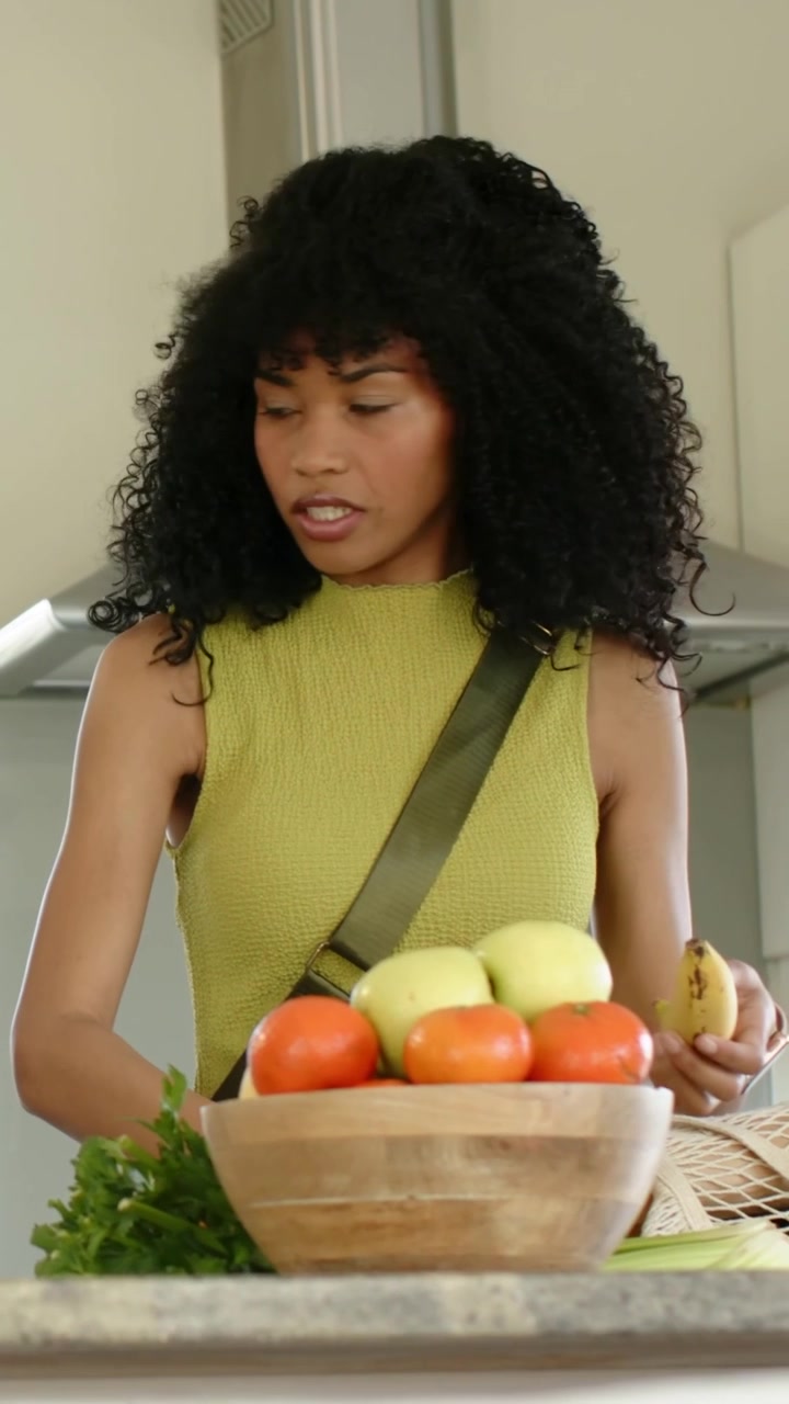 Video verticale: Donna con un top color oliva che sistema la spesa in cucina, mettendo il sedano e controllando una banana.