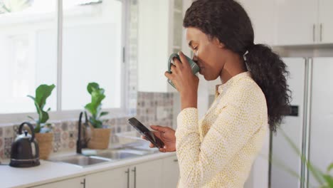 Video-Einer-Afroamerikanischen-Frau,-Die-Kaffee-Trinkt-Und-Ihr-Smartphone-In-Der-Küche-Benutzt