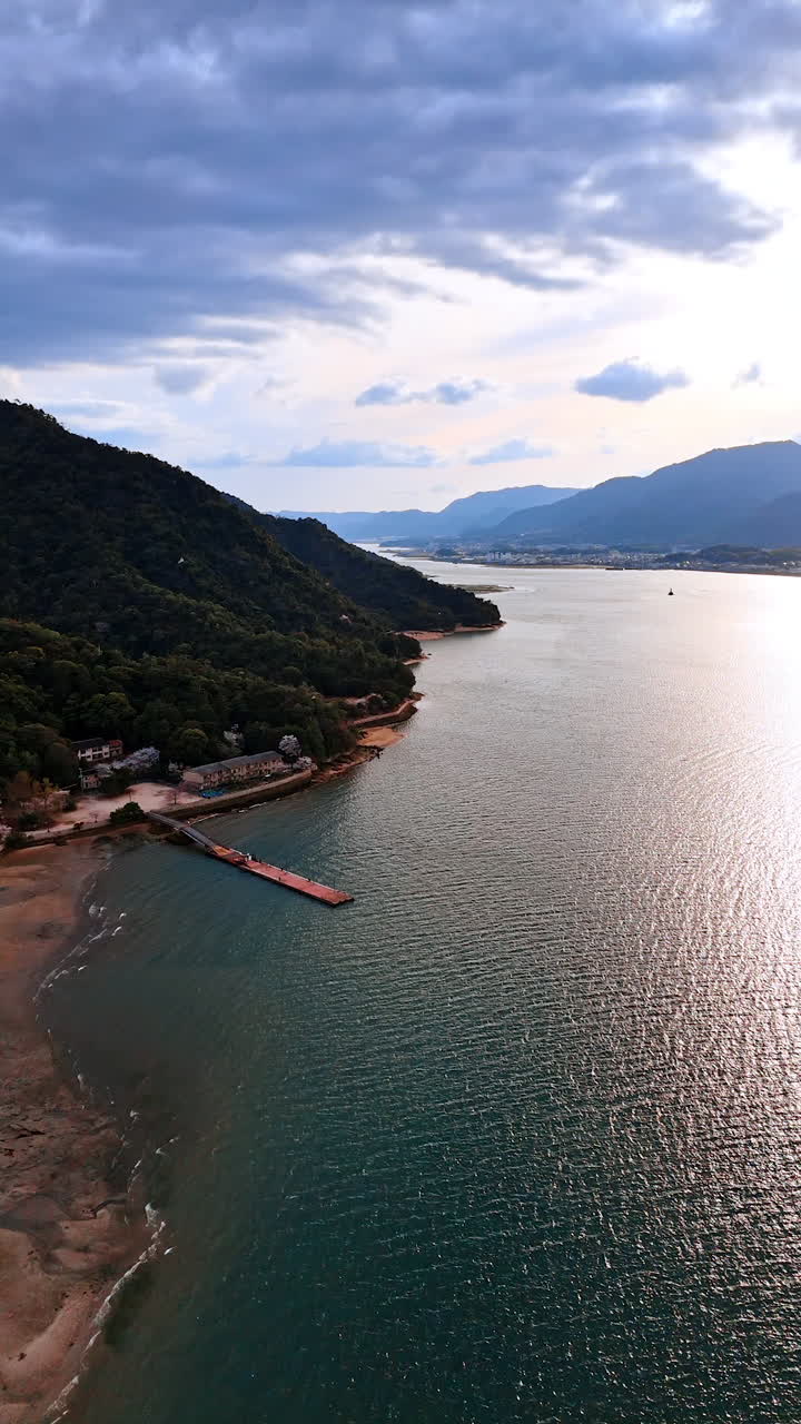 Splendid waterscape with ragged surface shining in the sun. Spectacular mountains covered with forests stand at the shores. Dramatic cloudscape covering the sky. Aerial view. Vertical video.