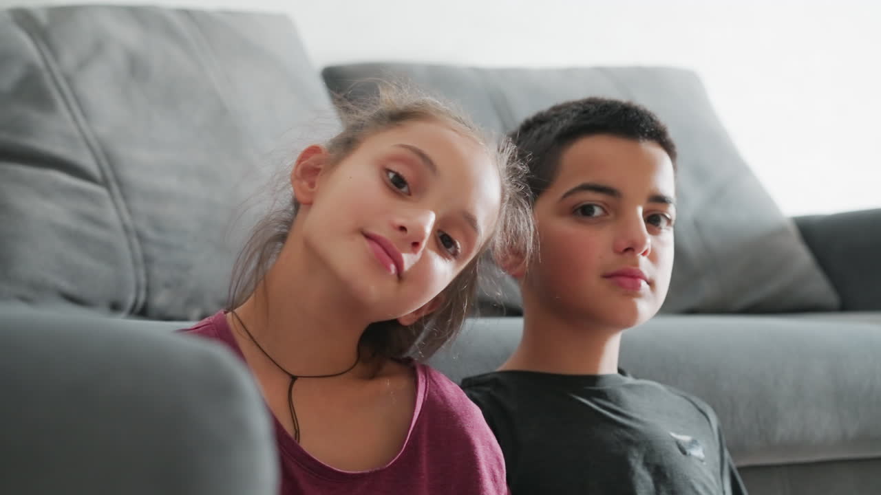 Una niña adorable apoya suavemente la cabeza en el hombro de su hermano mientras ambos se sientan juntos en el suelo del salón, apoyados en un sofá mullido, mirando a la cámara con expresiones sutiles que capturan el tierno vínculo fraternal.