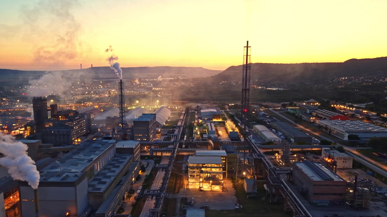 Arbeidere er travle i et stort industriområde ved solnedgang med lys og røyk som kommer fra fabrikker.