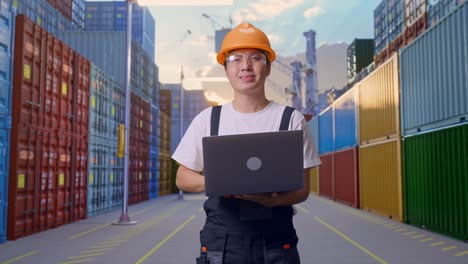 trabajador asiático con gafas de protección y casco de seguridad usando una computadora portátil y sonriendo mientras está de pie en el almacén del patio de contenedores