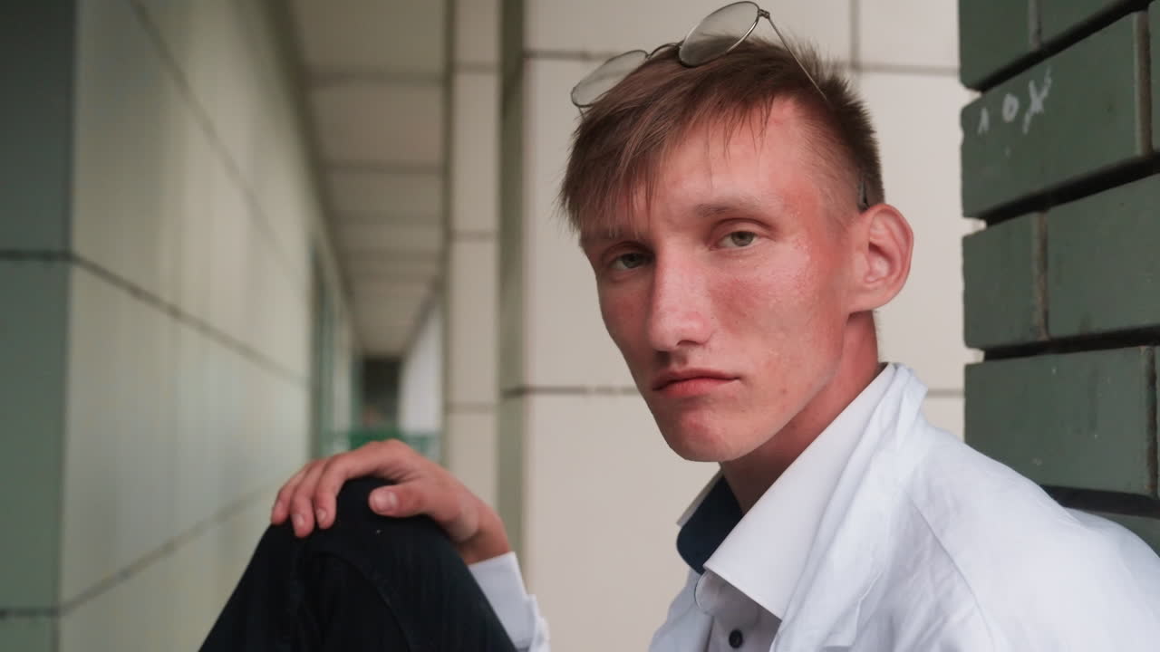 Young ecologist in white coat with glasses resting on head sits near wall gazing into distance with thoughtful expression, reflecting curiosity, observation, and scientific attention outdoors