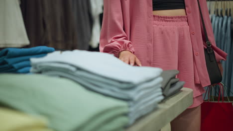 vista inferior de una dama con una camiseta negra debajo de una camisa rosa sin botones y pantalones cortos, llevando bolsas de compras, tocando suavemente la ropa bien plegada en una mesa mientras pasa por delante de ellos en una tienda minorista