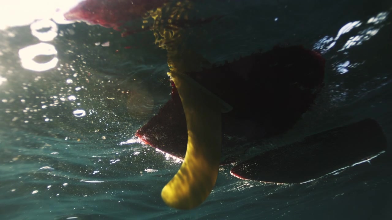 Surfboard fin rotates beneath shimmering ocean surface in dramatic upward underwater angle