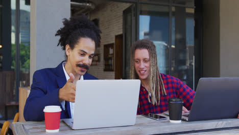 Two-diverse-male-friends-sitting-at-table-outside-cafe-drinking-coffee-and-using-laptop