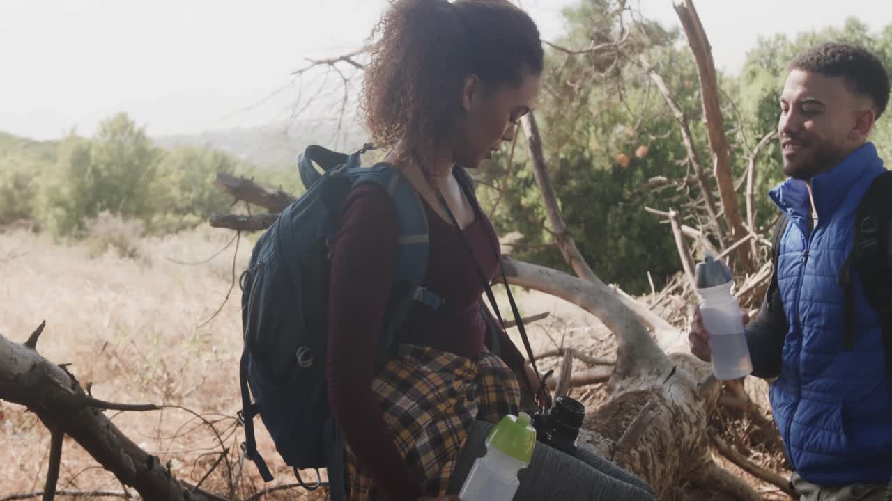 feliz pareja afroamericana sentada en el tronco de un árbol y bebiendo agua en el bosque, cámara lenta