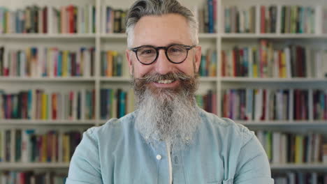 portrait of mature caucasian professor with beard smiling happy at camera in library bookshelf background wearing glasses