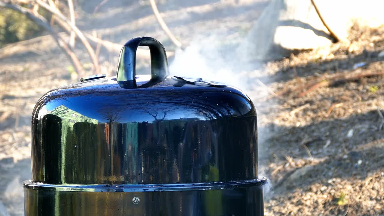 A person prepares a smoker for cooking meat in a backyard setting. Smoke rises as the smoker heats up under the sun in autumn. Friends enjoy the atmosphere around the smoker