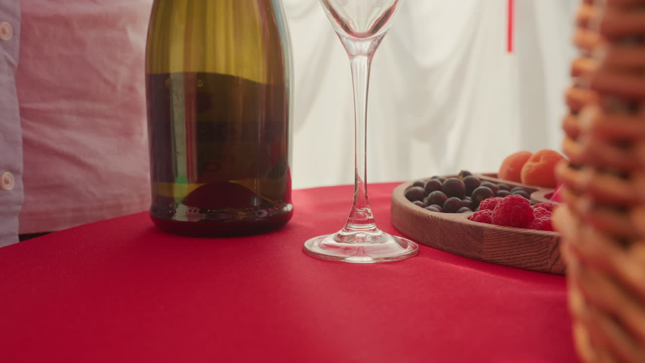 wine bottle on red tablecloth, hand reaches for stem glass beside wicker basket and tray of mixed berries, diffused sunlight through curtain, casual shirted partner arranging intimate date scene