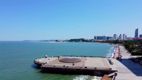 slow aerial forward shot of the coast from beautiful xingfu park in weihai, china with a view to the blue sea and city with skyscrapers in background and pedestrians on a platform on sunny day