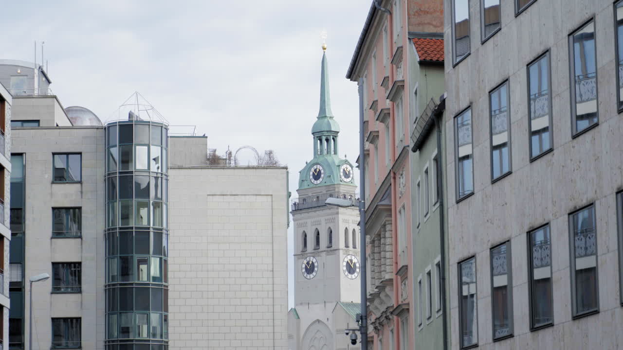 A historic clock tower with a green spire rises between modern and classic buildings in an urban European setting. The mix of architectural styles reflects the city's rich history and development.