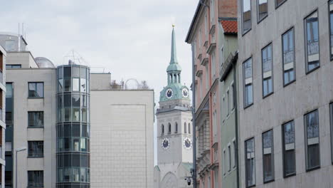 a historic clock tower with a green spire rises between modern and classic buildings in an urban european setting. the mix of architectural styles reflects the city's rich history and development.