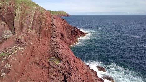 Drone-ariel-footage-of-red-Welsh-cliffs-with-blue-sky's-and-crashing-waves