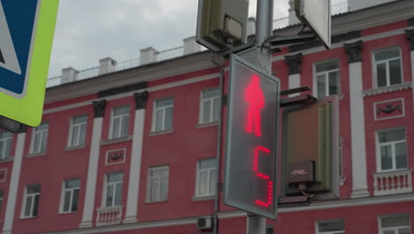 close view of a pedestrian crossing sign post with a neon yellow border and a digital traffic light showing a countdown. a red-colored city building background under a cloudy sky