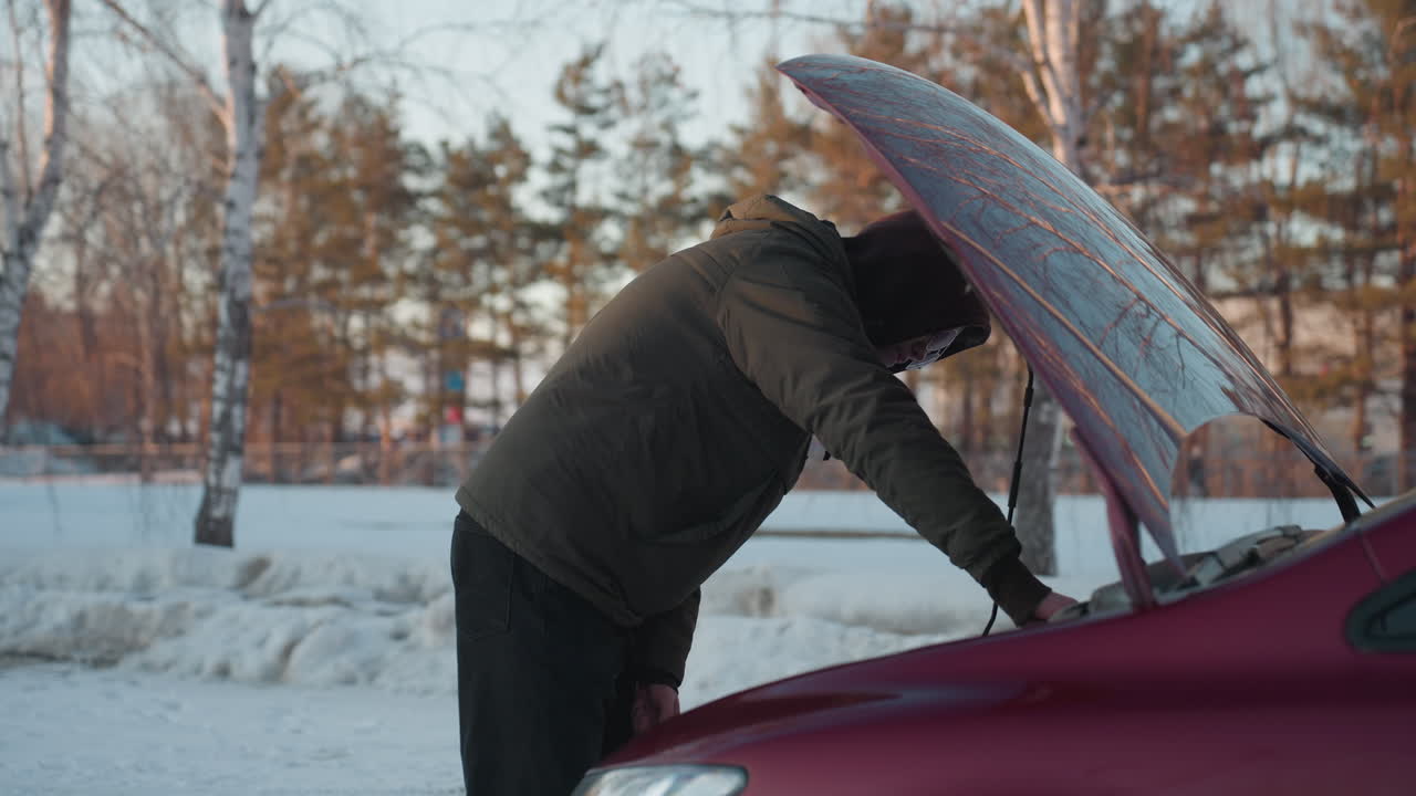 Young man wearing hoodie inspects oil level using dipstick while standing beside parked red car with bonnet open in cold outdoor environment surrounded by snow and bare trees in winter