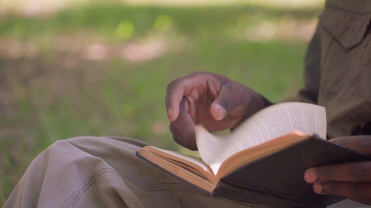 Close up of person reading open book outdoors with their hand turning page. person is seated in nature wearing casual clothing focusing on text visible through clear daylight lighting