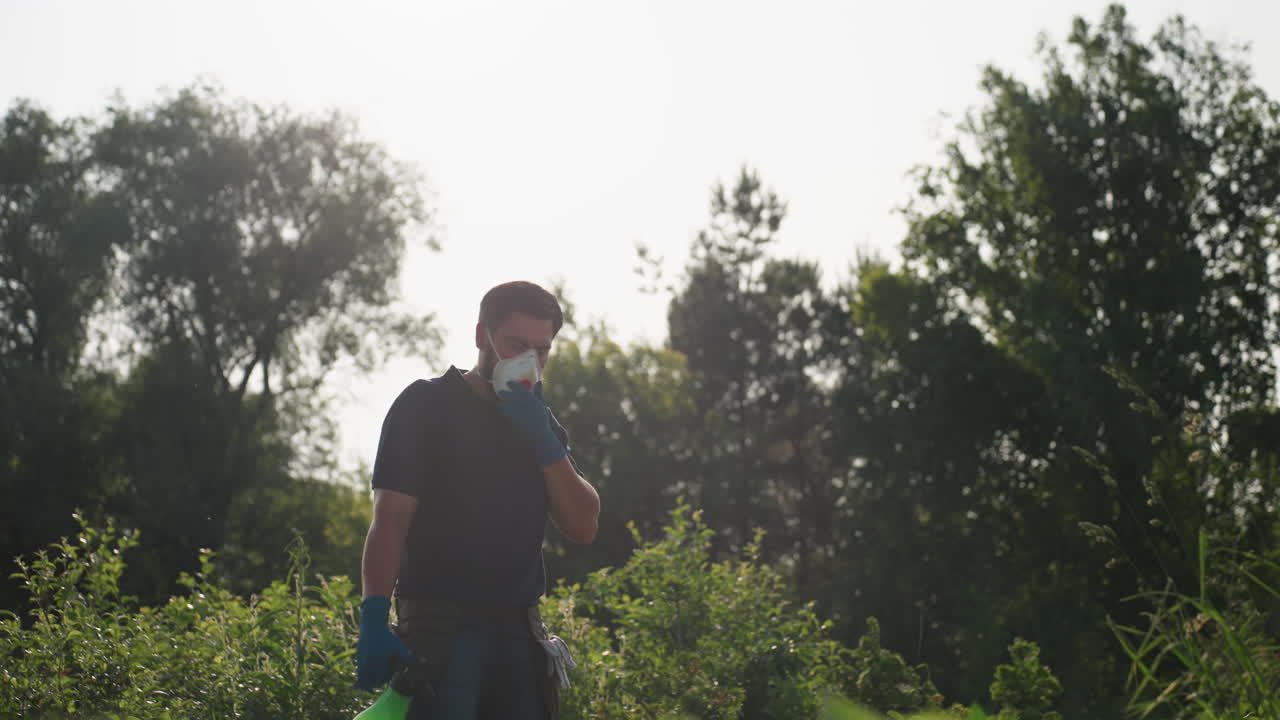 Male gardener wearing protective mask and gloves adjusts nose area of mask while preparing to fumigate plants in lush green outdoor environment under bright sunlight