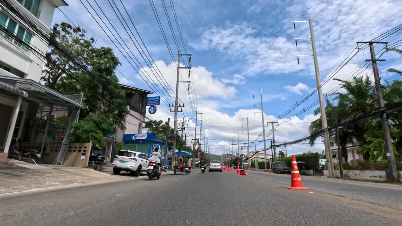 Vehicle moves down sunlit Phuket street, passing motorcycles, cars, and orange traffic cones