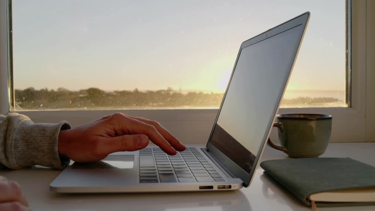 Iført grå sweatshirt åbner en person sin bærbare computer ved skrivebordet ved vinduet og begynder at arbejde med en grøn kop og notesbog.